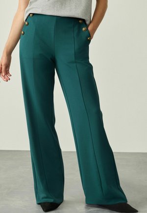 Grüne weit geschnittene Hose aus glattem Stoff, mit goldenen Knopfakzenten an der Vorderseite und Seitentaschen. Feine Streifendetails sind sichtbar.