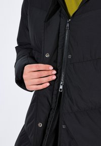 Černá puffer bunda se zapínáním na zip, patentkami a vysokým límcem. Materiál má hladkou texturu s prošívaným vzorem.