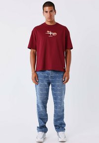 Modelo masculino vistiendo una camiseta de color burdeos con diseño floral, jeans azules con un patrón de escritura blanca, zapatillas blancas, de pie contra un fondo liso.