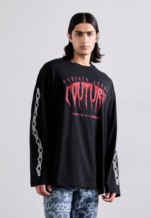 Jeune homme avec des cheveux mi-longs portant une shirt à manches longues noir avec le texte "Couture" en rouge et des motifs de chaînes sur les manches, ainsi que des jeans à motifs.