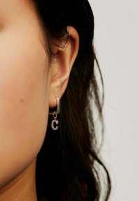 Pendiente de aro de plata con un charm en forma de letra "C" estilizada, que muestra una textura suave y adornos brillantes, llevado en la oreja.