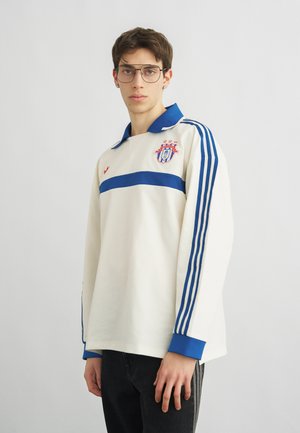 Νεαρός άνδρας με γυαλιά που φοράει ένα λευκό αθλητικό μπλουζάκι μακρυμάνικο με μπλε ρίγες και έμβλημα ομάδας στο στήθος.