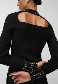 Femme portant un haut noir à manches longues avec un dos ouvert et des ornements en strass sur le col et les poignets, les mains croisées derrière le dos.