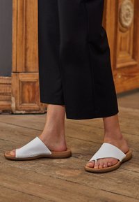White leather slide sandals with a flat brown sole, featuring a wide strap and decorative stitching along the edge.