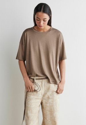 Kobieta w luźnej brązowej bluzce z krótkim rękawem i jasnobeżowych spodniach w wzory, regulująca sznurek w spodniach, stojąca na tle gładkiej białej ściany.