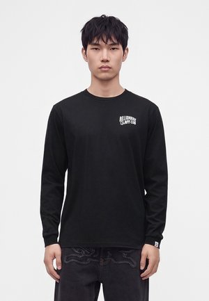 Ung mand med sort hår har en sort langærmet skjorte med "Billionaire Boys Club" logo på brystet og mørke jeans med broderi.