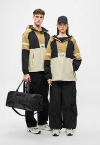 Μπεζ και μαύρα μπουφάν με κουκούλα, φερμουάρ και λογότυπο· μαύροι παντελόνια cargo· μαύρη τσάντα duffel· αθλητικά παπούτσια. Άνδρας και γυναίκα μοντέλα ποζάρουν μαζί.