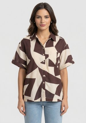 Vrouw met donker golvend haar die een kort mouwens overhemd draagt met abstracte bruine en beige patronen en lichtblauwe spijkerbroek.