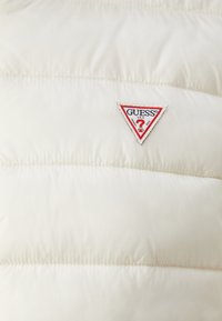 Vitt vadderat väst med ett broderat mönster och en triangulär röd, vit och svart "GUESS" logotipp på framsidan. Slät textur.