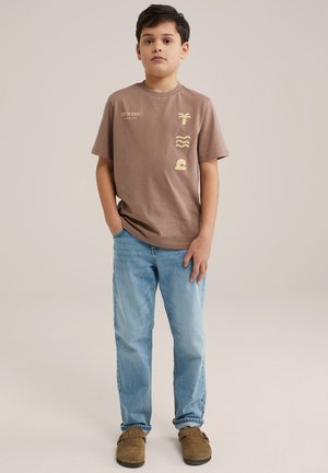 T-shirt marron à manches courtes avec des graphiques jaunes : un palmier, des lignes ondulées et un design de vagues. Associé à un jean bleu clair et des chaussures marron.