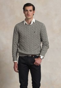 Suéter gris de punto trenzado con cuello redondo, llevado sobre una camisa blanca de cuello, combinado con jeans oscuros y un reloj plateado.