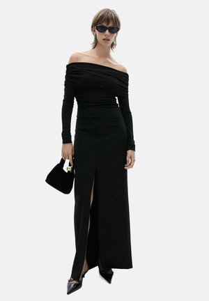 Black off-shoulder dress with long sleeves, fitted design, and side slit. Worn with black sunglasses and a small black handbag, featuring pointed toe heels.