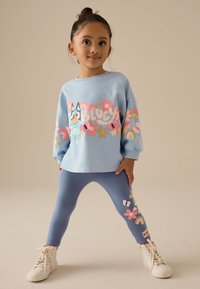 Sudadera azul claro con gráfico divertido, combinada con leggings azules con patrones florales. Zapatillas blancas de caña alta completan el conjunto.