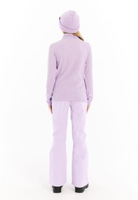 Moteris, dėvinti levandų spalvos megztą kepurę, įridytą golfo tipo megztinį ir sniego kelnes, stovi atsukusi nugarą baltoje fone.