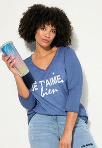 Camisa azul de manga larga con escote en V y texto blanco "JE T'AIME BIEN", acompañada de un vaso de arcoíris en degradado con una superficie texturizada.
