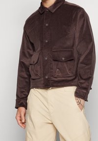 Brun manchesterjacka med framknäppning, spetsig krage och två framfickor. Slät textur och kort design.