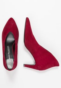 Zapatos de tacón alto de ante rojo con punta afilada, diseño ajustado y un elegante tacón de aguja. Forro negro y detalles de costura sutiles visibles.