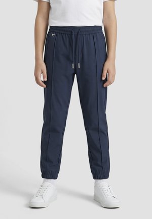 Persona que lleva pantalones jogger azul marino con puños elásticos, detalles en la costura frontal, cintura con cordón, zapatillas blancas y una camiseta blanca.