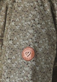 Blommönstrad tyg i nyanser av grönt och brunt med en texturerad yta. Har ett bronsmärke med ett hjärtmotiv.