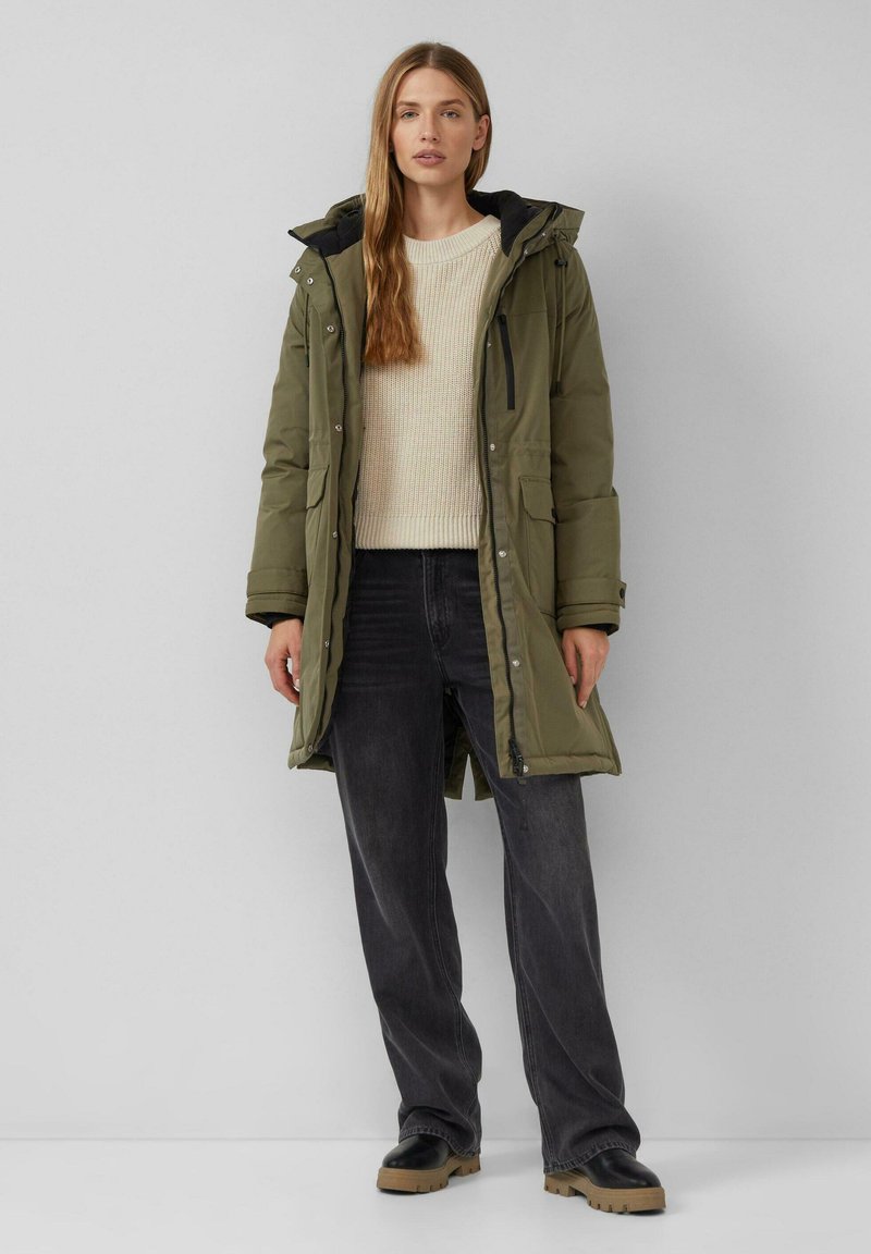 Olijfgroene parka met een rits- en knoopsluiting, capuchon, voorkruisen en zwarte voering, gedragen over een crèmekleurige gebreide trui en donkergrijze broek.