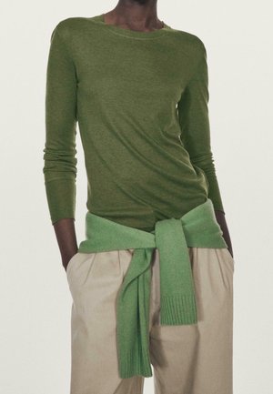 Person iført en olivengrøn, langærmet skjorte og beige bukser med en lysegrøn sweater bundet omkring taljen, hænderne i lommerne.