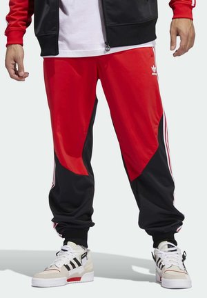 Osoba ubrana w czerwono-czarne spodnie dresowe Adidas i białe buty sportowe Adidas z czarnymi paskami, stojąca na jednolitym tle.