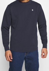 Mörkblå långärmad tröja i sweatshirtmaterial, med rund halsringning och en liten logotyp på vänster bröst.