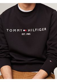 Schwarzer Pullover mit weißem, gesticktem Schriftzug "TOMMY HILFIGER EST. 1985" und einem dreifarbigen Logo, mit gerippten Bündchen und Rundhalsausschnitt.