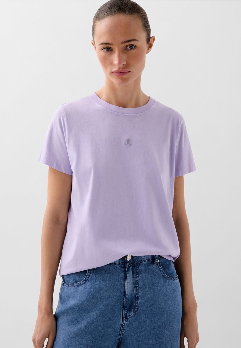 T-shirt in cotone lavanda con colletto rotondo e maniche corte. Presenta un discreto dettaglio del logo sul petto. Abbinato a una gonna in denim blu.