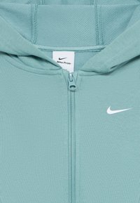 Haut de survêtement zippé Nike bleu clair avec capuche, fermeture éclair visible et logo Nike swoosh blanc sur la poitrine.