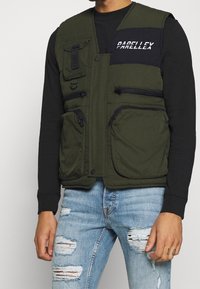 Grüne gesteppte Weste mit mehreren Taschen, Reißverschlüssen und einem Logo-Patch auf der Brust; getragen über einem schwarzen Langarmshirt und blauen, zerfetzten Jeans.