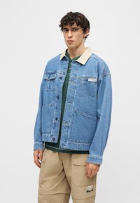 Νέος άνδρας με κατσαρά μαλλιά και γυαλιά, φορώντας μπλε τζιν μπουφάν πάνω από πράσινο πουκάμισο και μπεζ παντελόνι cargo, σε απλό φόντο.