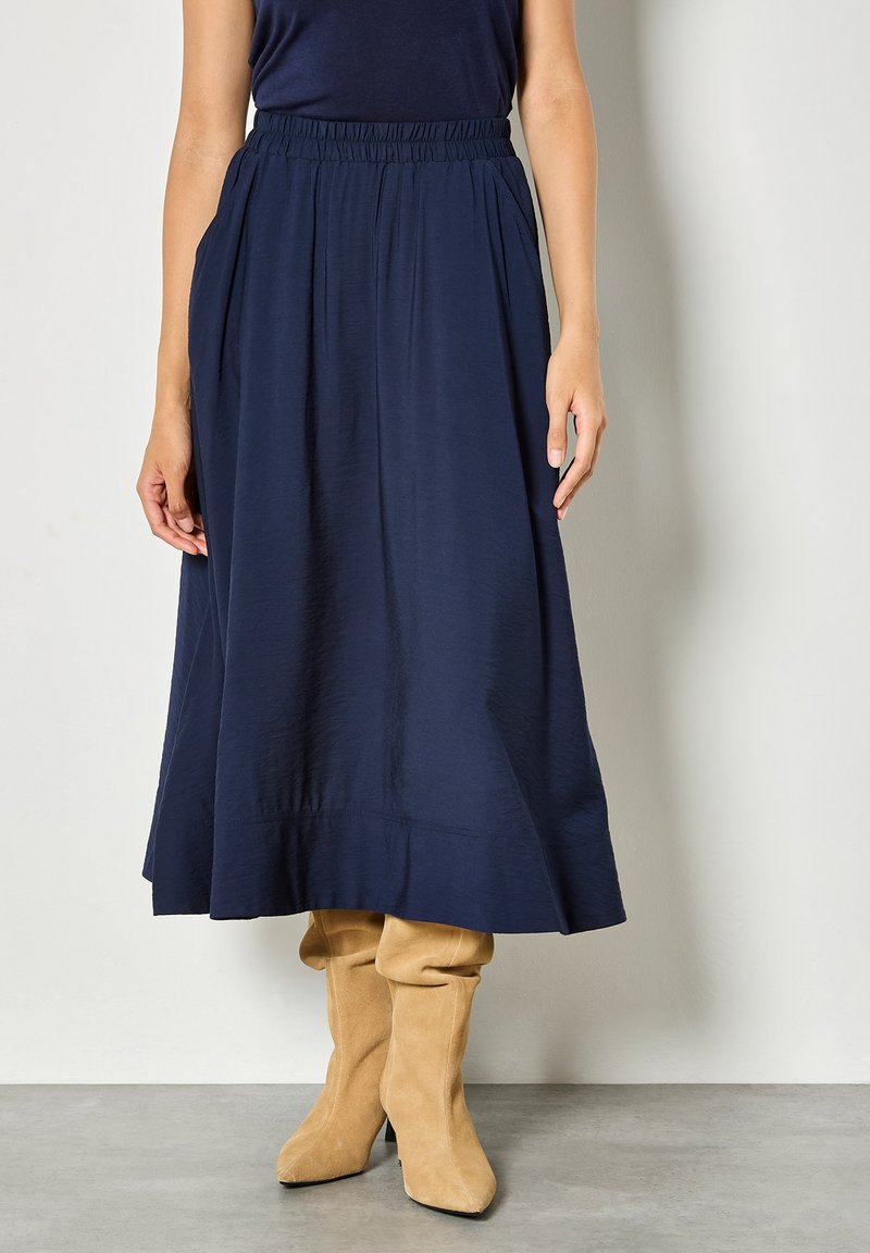 Personne portant une jupe midi bleu marine avec une taille élastique et des bottes hautes jusqu'aux genoux en daim marron clair, debout sur un sol gris clair.