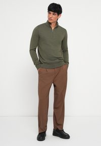 Olive green zip-neck sweater with a ribbed hem, paired with brown tailored trousers and black sneakers. Simple, textured knit design.