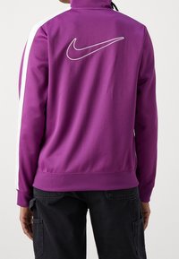 Person som bär en lila Nike träningsjacka med vita ränder på ärmarna och en stor vit Nike-swoosh på ryggen, kombinerad med svarta byxor.