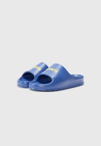 Blue rubber slide sandals with an open toe design. Features raised text in yellow reading "LACOSTE" on the upper. Textured sole for grip.