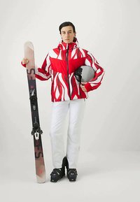 Giacca da sci a motivi rossi e bianchi, pantaloni bianchi, scarponi da sci neri, che tiene gli sci con attacchi e un casco nero.