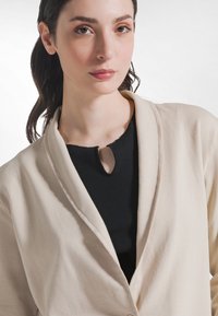 Lungo giacca beige con collo sciallato, caratterizzata da una texture liscia e un'apertura frontale aperta, indossata sopra una maglia nera a costine con scollo a goccia.