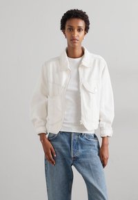 Balts īsais džersija ar lielām kabatām, redzamām šuvēm un apkakli, uzvilkts virs baltas blūzes, savienots ar zilām džinsām.