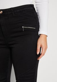 Vaqueros negros con un bolsillo con cremallera y paneles texturizados en los muslos, combinados con una camiseta blanca de manga larga. Vista en primer plano de la zona de la cintura.