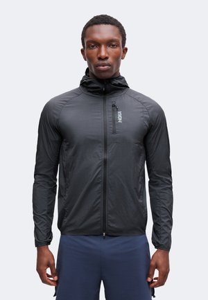 Homme portant une veste à capuche noire ajustée avec une petite poche poitrine et un short de sport bleu marine, debout devant un fond uni.