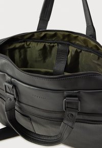 Fekete bőrtáska sima és texturált részekkel, zöld belső béléssel, cipzáras záródással és dupla fogantyúval. Kontrasztos díszítésekkel rendelkezik.