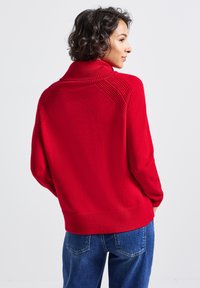 Frau mit kurzem lockigem Haar, die einen roten Strickpullover mit strukturierten Schulterpartien und blauen Jeans trägt, steht vor einem schlichten weißen Hintergrund.