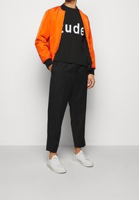 Oranssi pommitakki, musta t-paita valkoisella tekstillä, mustat kapenevat housut ja valkoiset tennarit. Yksinkertainen, moderni muotoilu ja värikontrasti.