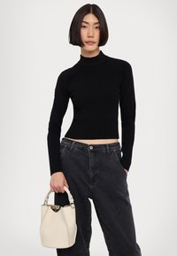 Svart ribbad polotröja med långa ärmar, kort design. Modellen håller en beige bucket bag med en texturerad yta och ett kort handtag.