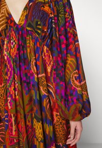 Vibrant langærmet kjole med abstrakt blomster- og dyreprint i røde, orange, lilla, grønne og blå nuancer, båret af en person med hånden synlig.