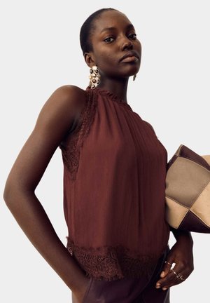 Femme portant un haut en dentelle marron sans manches, de grandes boucles d'oreilles dorées, tenant un sac patchwork beige et marron, avec des cheveux courts et une expression neutre.