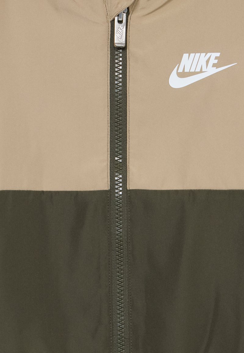 Nike-Jacke mit einem beigen Oberteil und einem dunkelgrünen Unterteil, ausgestattet mit einem Metallreißverschluss und einem weißen Logo auf der Brust.