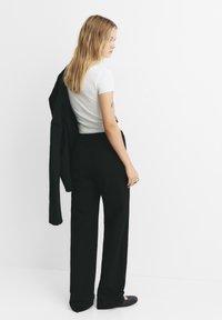 Ung kvinna med långt blont hår som bär en vit t-shirt och svarta byxor, hållande en svart jacka över axeln, står sidledes.