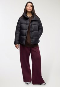 Młoda kobieta ubrana w czarną kurtkę puchową, brązowy sweter, szerokie burgundowe spodnie i białe trampki, stojąca na tle jednolitym.